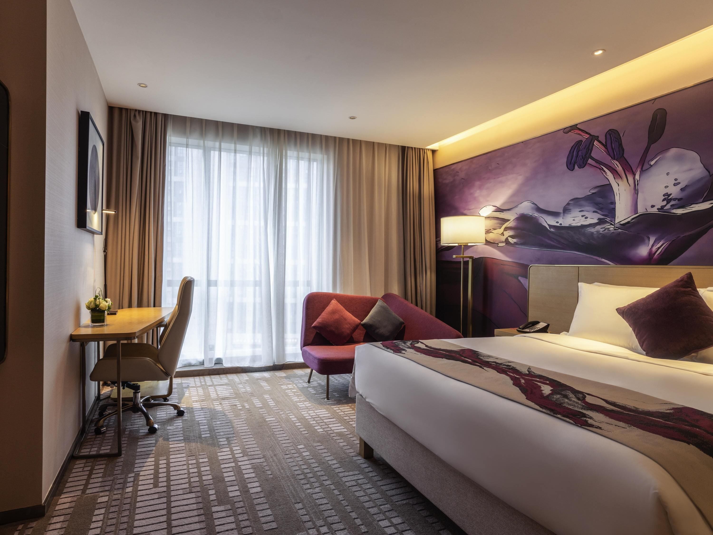 A hotel room with a bed, a desk, a chair, a red armchair, a painting on the wall, and a window with curtains.