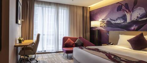 A hotel room with a bed, a desk, a chair, a red armchair, a painting on the wall, and a window with curtains.