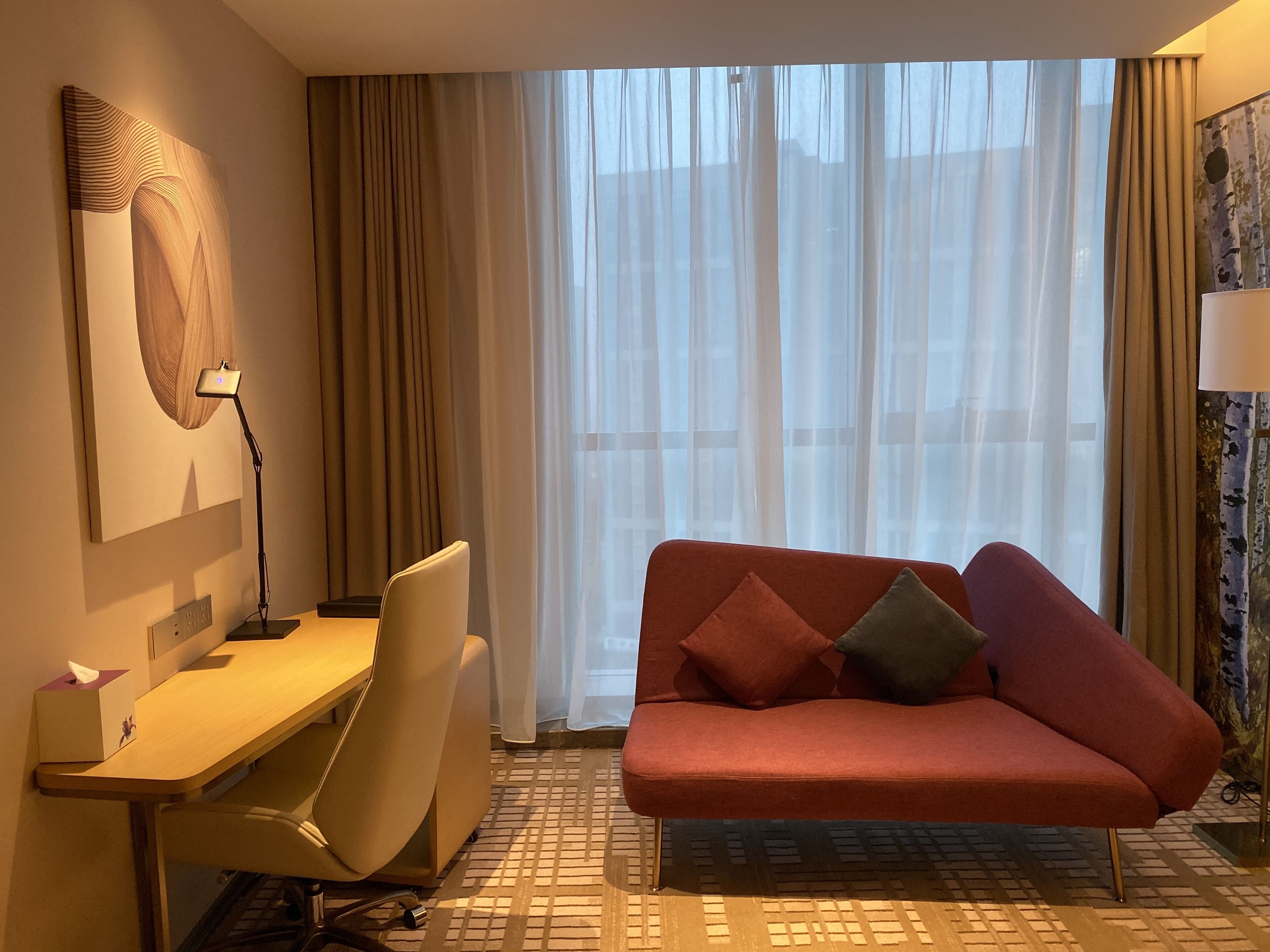 Une chambre d’hôtel avec un bureau, un canapé rouge, une lampe et une grande fenêtre ornée de rideaux légers.