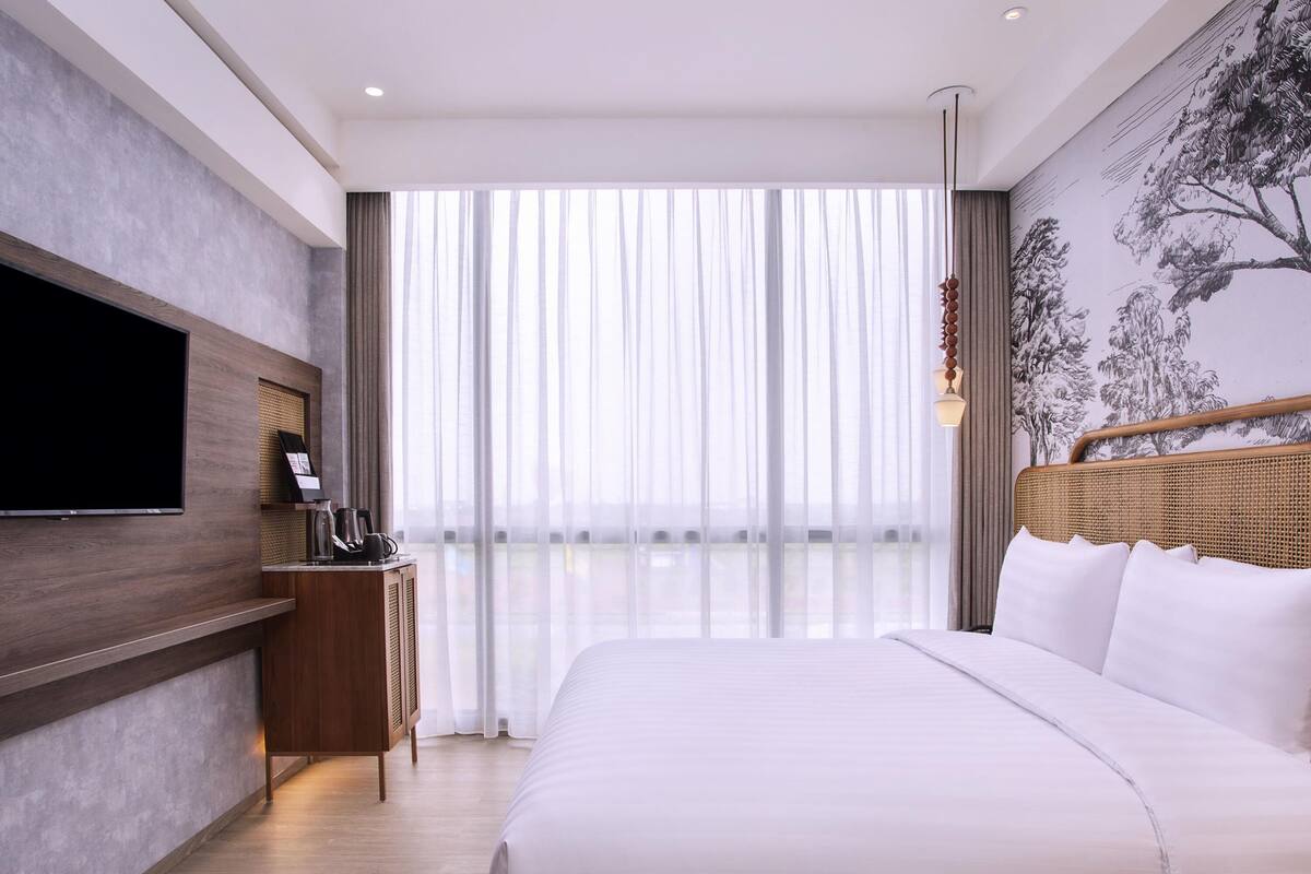 Ein modernes Hotelzimmer mit einem großen Bett, einem Flachbildfernseher, einer Kaffeemaschine und Blick ins Freie durch transparente Vorhänge.