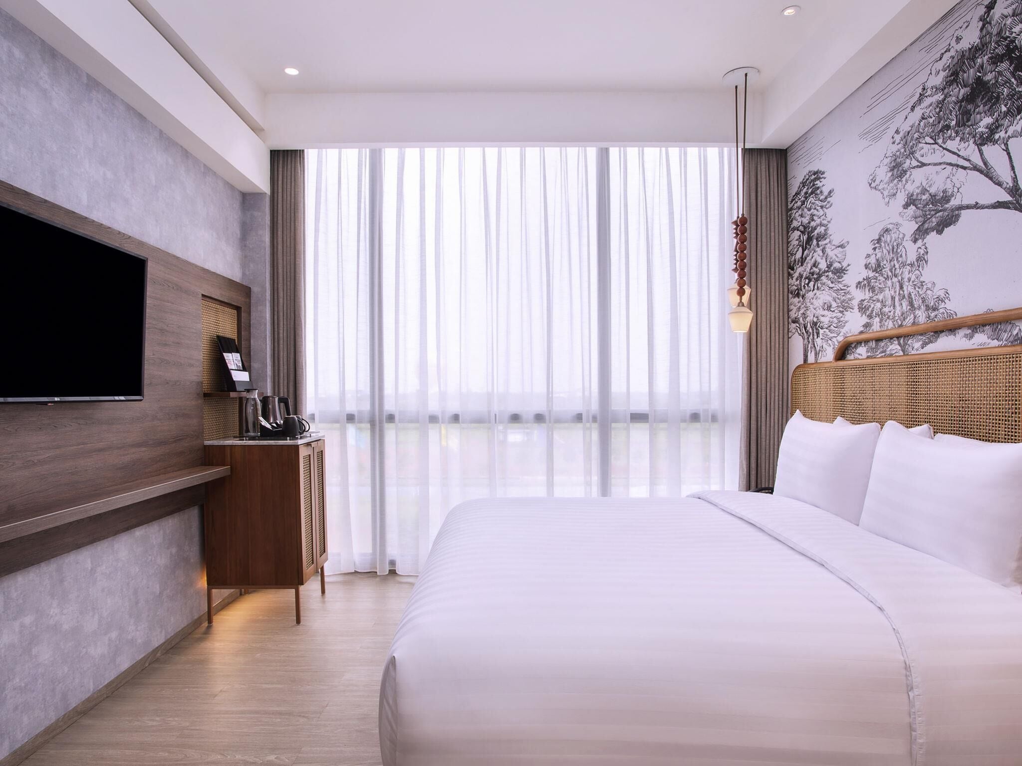 A modern hotel room with a large bed, a flat-screen TV, a small side table with a coffee maker, and a large window with sheer curtains.
