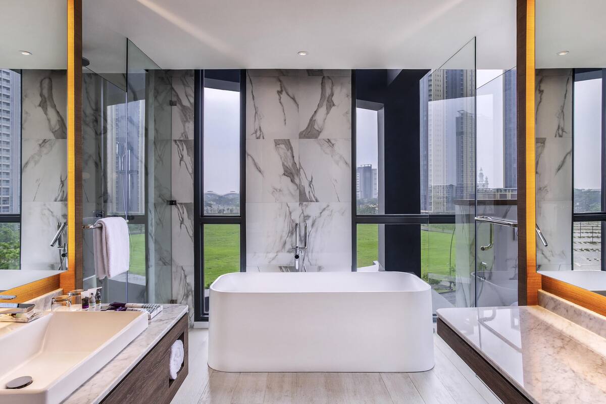 Une salle de bain moderne dotée d’une grande baignoire, de murs en marbre et de grandes fenêtres offrant une vue sur la ville.