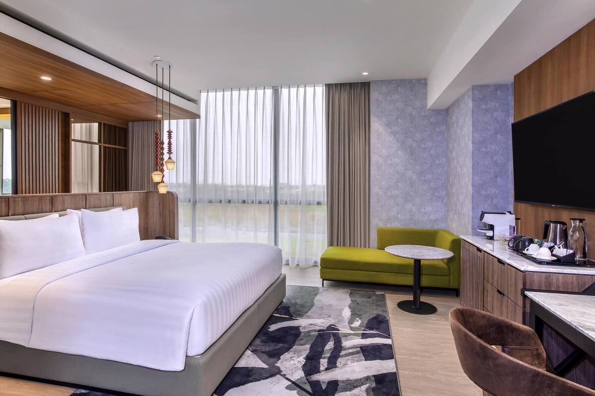 Ein modernes Hotelzimmer mit einem großen Bett, einem grünen Sessel, einem kleinen runden Tisch, einem Schreibtisch mit einem Flachbildfernseher und einer Kaffeestation.