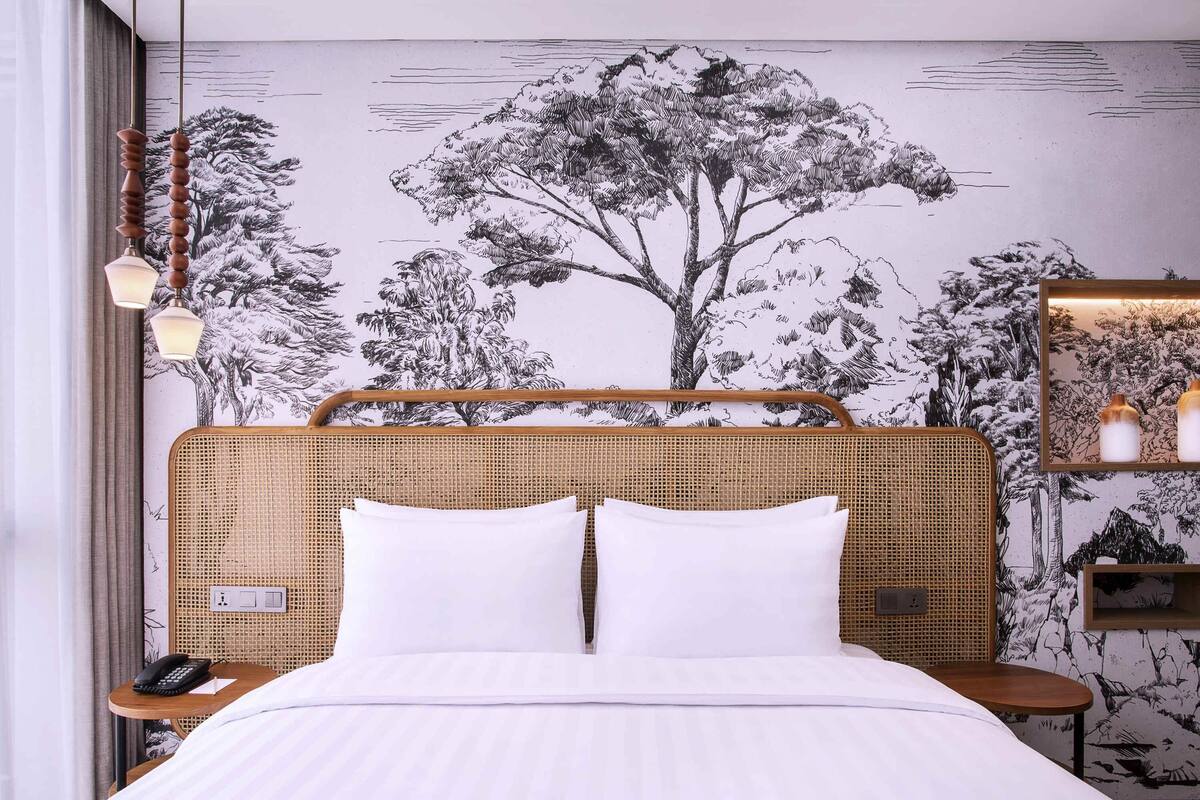 Un lit bien fait, avec du linge de lit blanc, une tête de lit en osier et une fresque murale représentant des arbres.