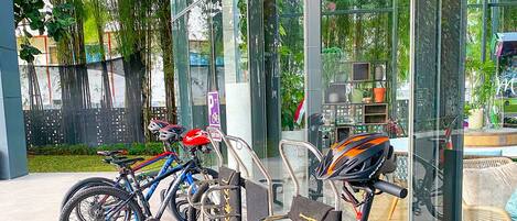 A row of bicycles and scooters parked outside a building with glass walls, reflecting greenery and a clear sky.