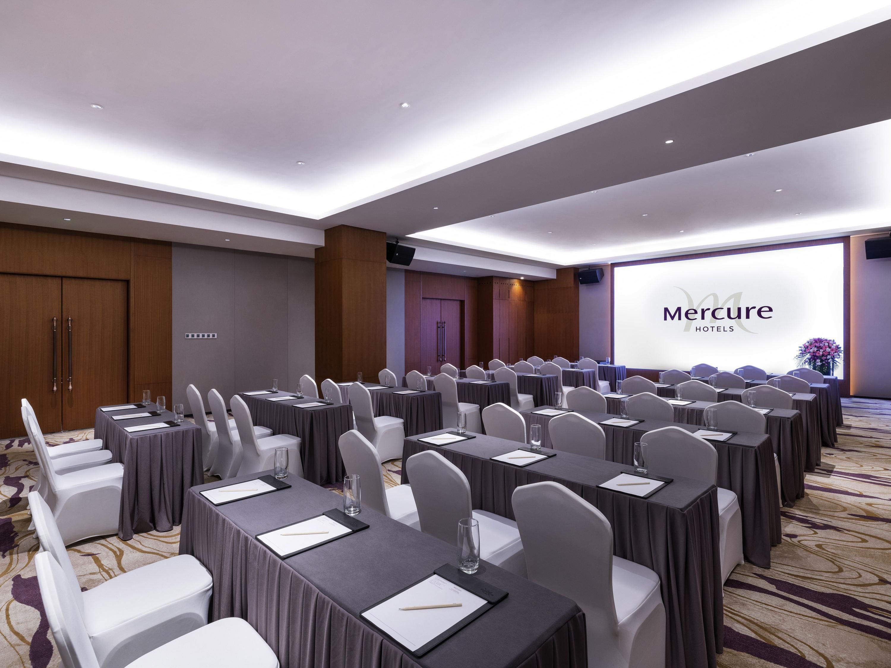 A conference room with rows of tables and chairs, a large screen displaying 'Mercure Hotels', and a wooden door in the background.