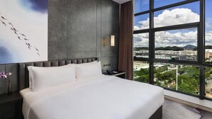 A modern hotel room with a large bed, a view of the city through floor-to-ceiling windows, a painting on the wall, and a nightstand with a lamp. - Shenzhen Longgang Kunyi Mercure Hotel (Shenzhen)