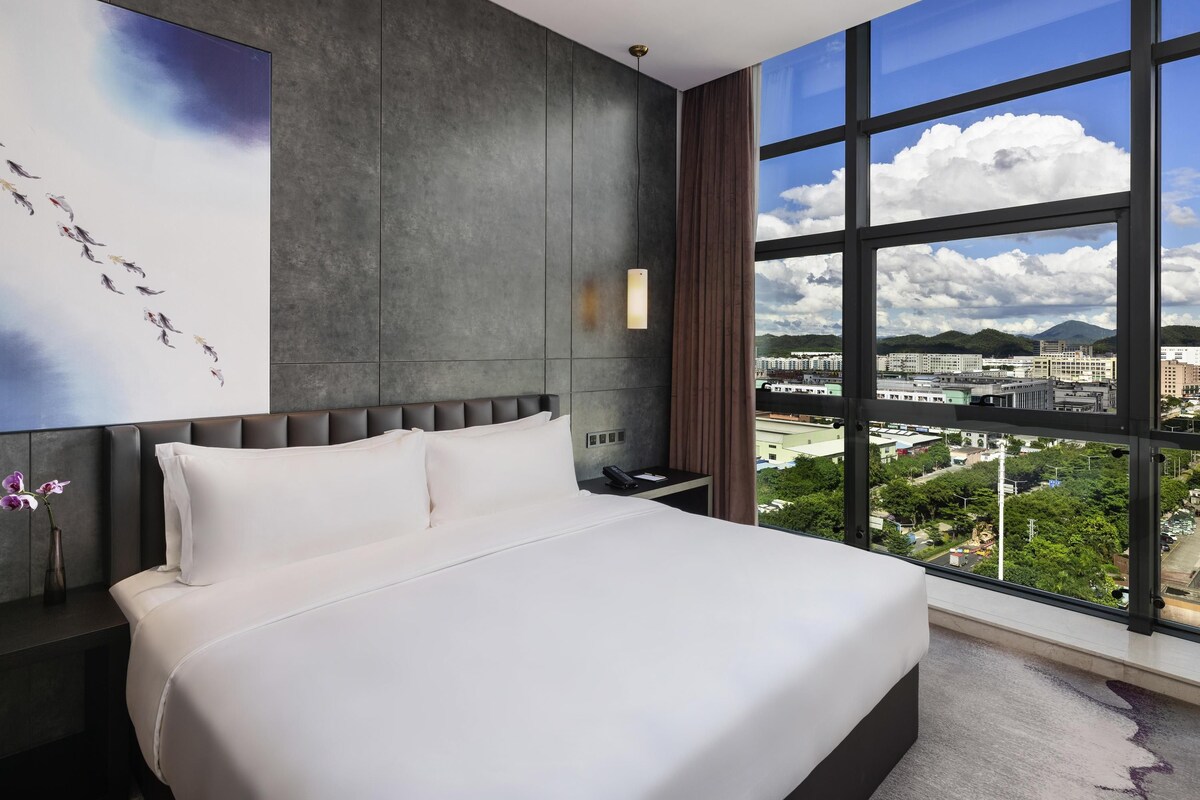 A modern hotel room with a large bed, a nightstand, a wall-mounted lamp, and a view of a cityscape through large windows.