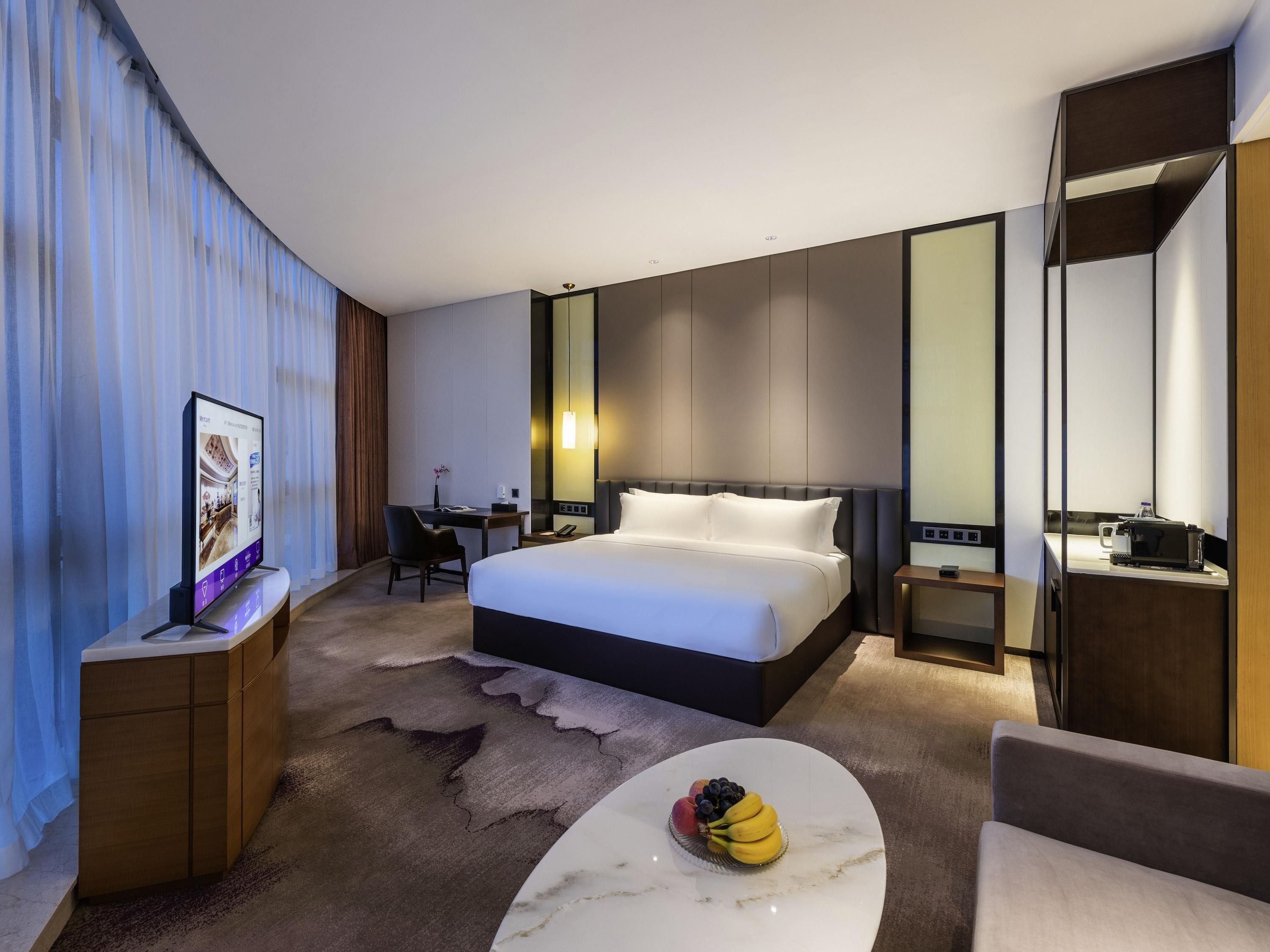 A modern hotel room with a large bed, a desk with a computer, a small round table with fruit, and a large mirror.
