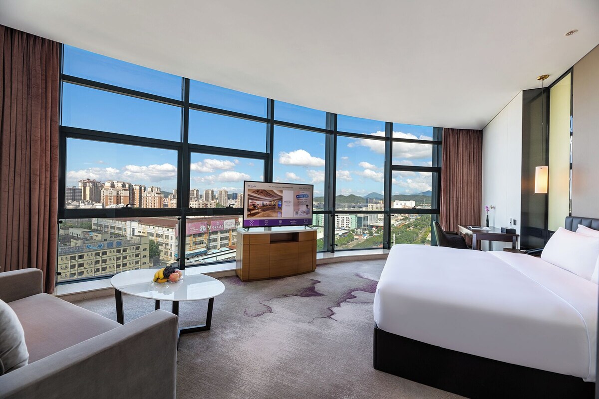 A hotel room with a large bed, a desk, a chair, a small table with fruit, and a TV.