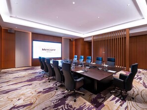 Uma sala de conferência com uma mesa grande, várias cadeiras, uma tela de projetor exibindo 'Mercure Hotels' e um tapete estampado.