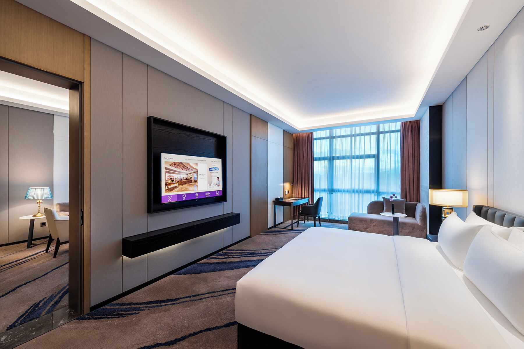 A hotel room with a large bed, a desk, a chair, a TV mounted on the wall, and a window with curtains.