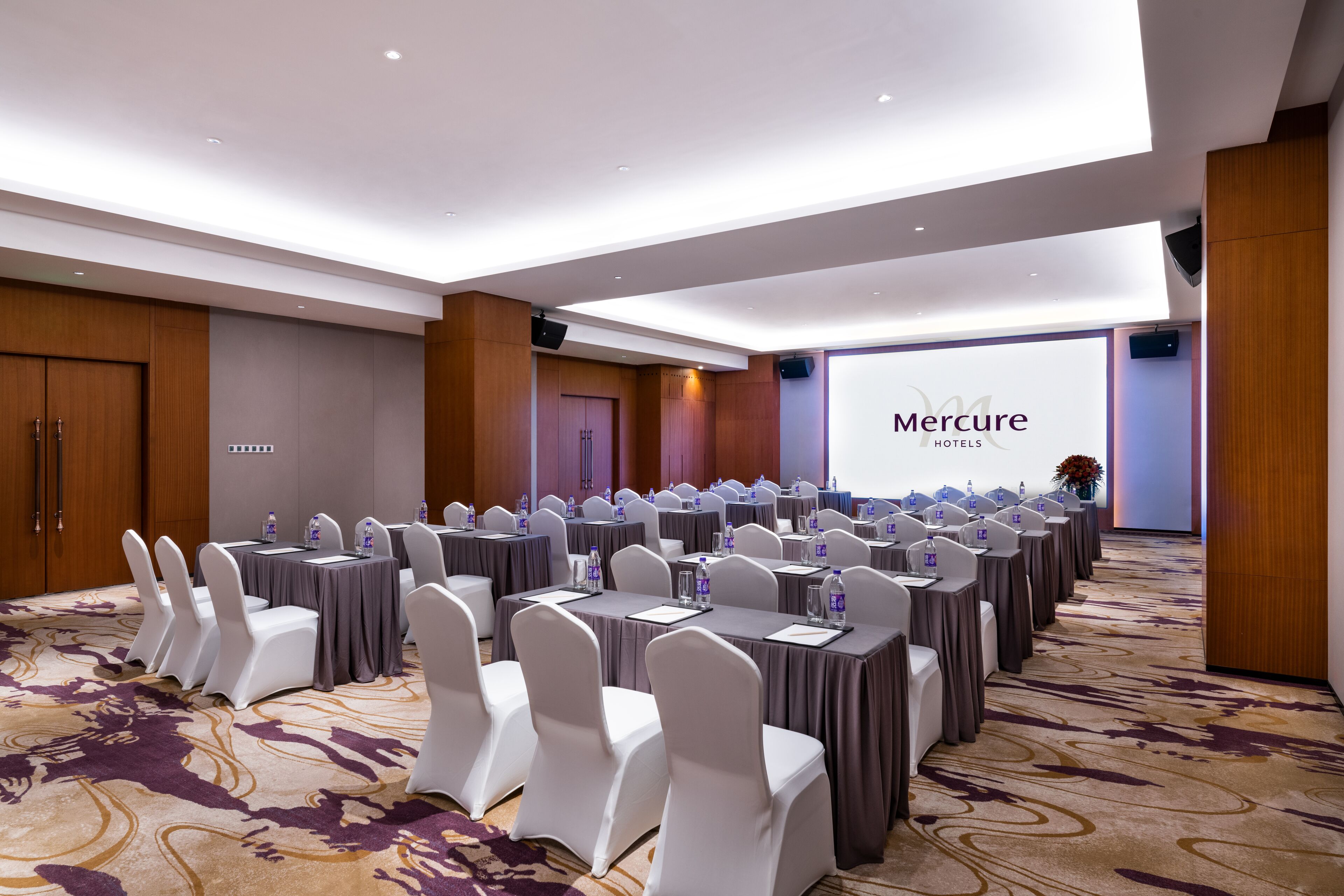 A conference room with rows of tables and chairs, a large screen displaying 'Mercure Hotels', and a carpeted floor.