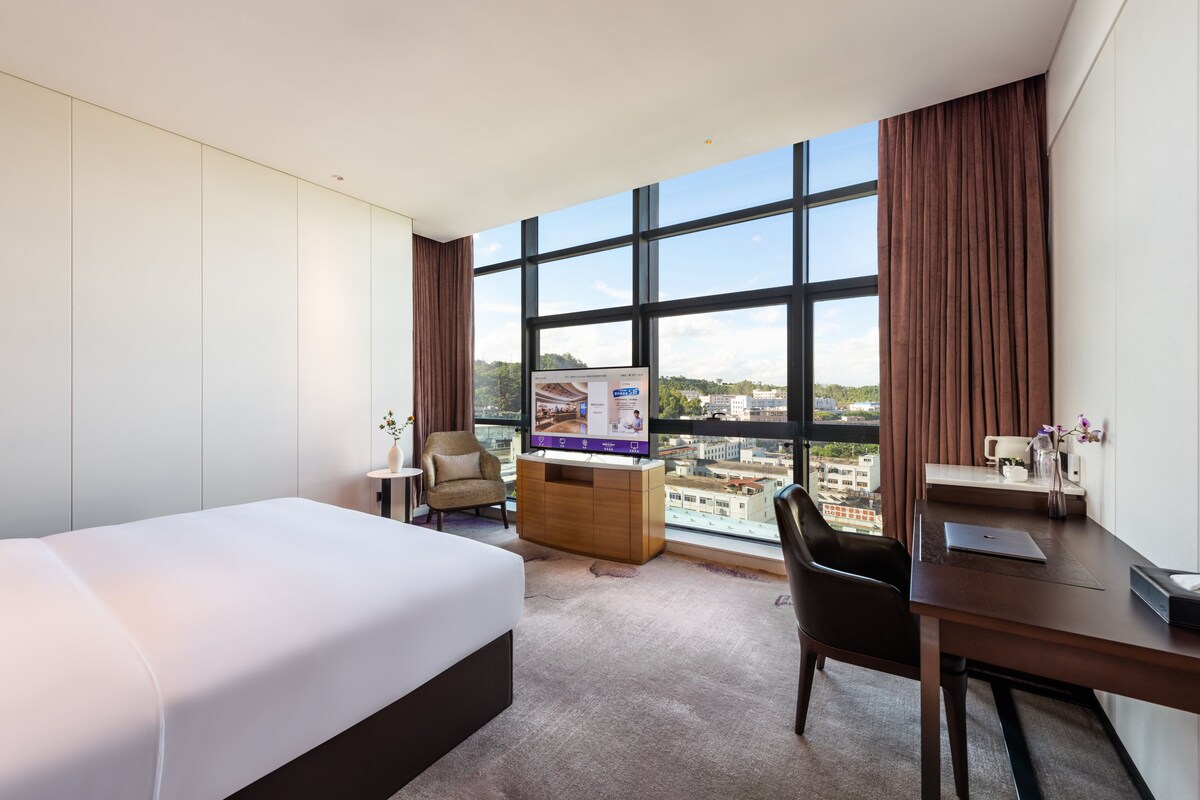A hotel room with a bed, a desk, a chair, a TV, and a large window with a city view.
