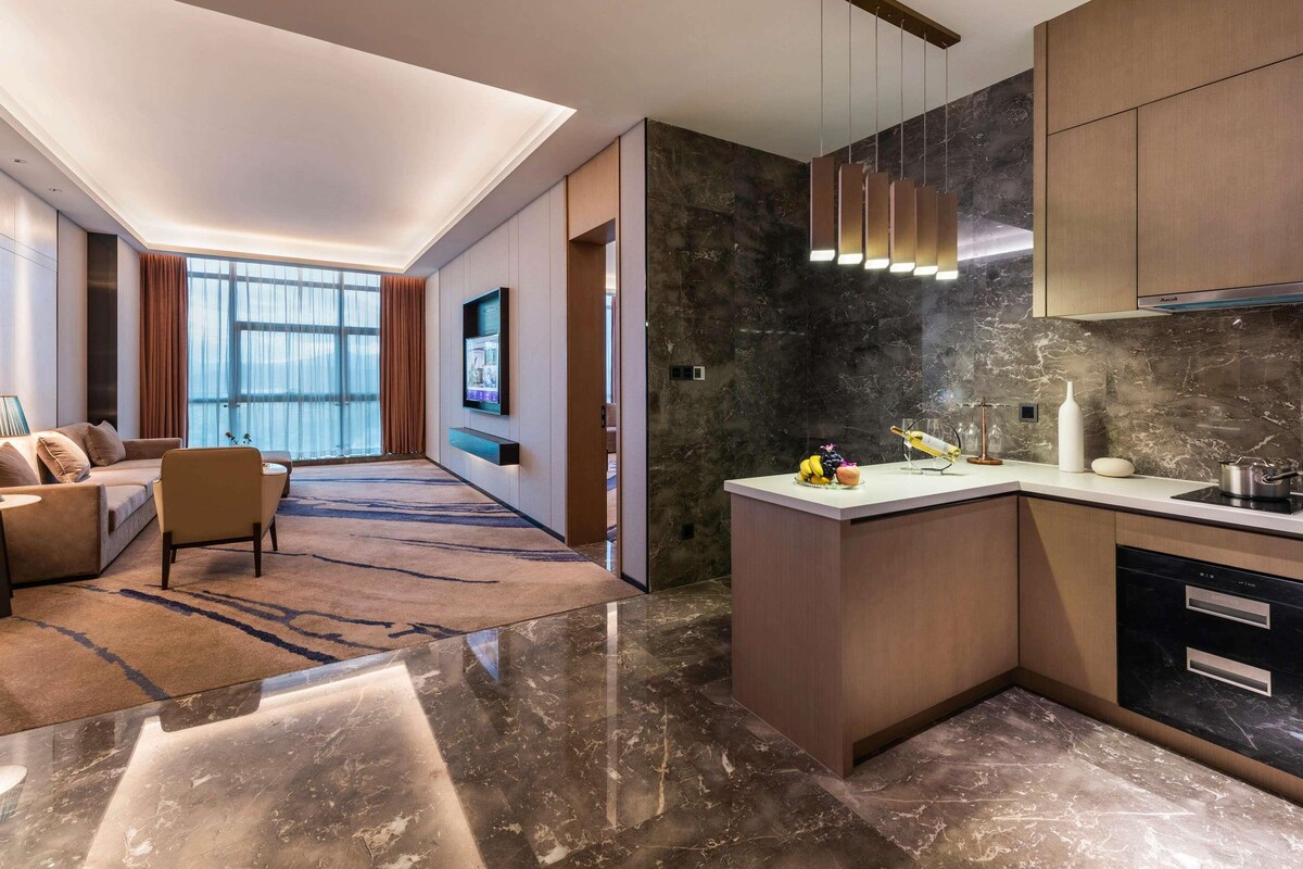 A modern hotel room with a living area, a kitchen, and a large window offering a view.