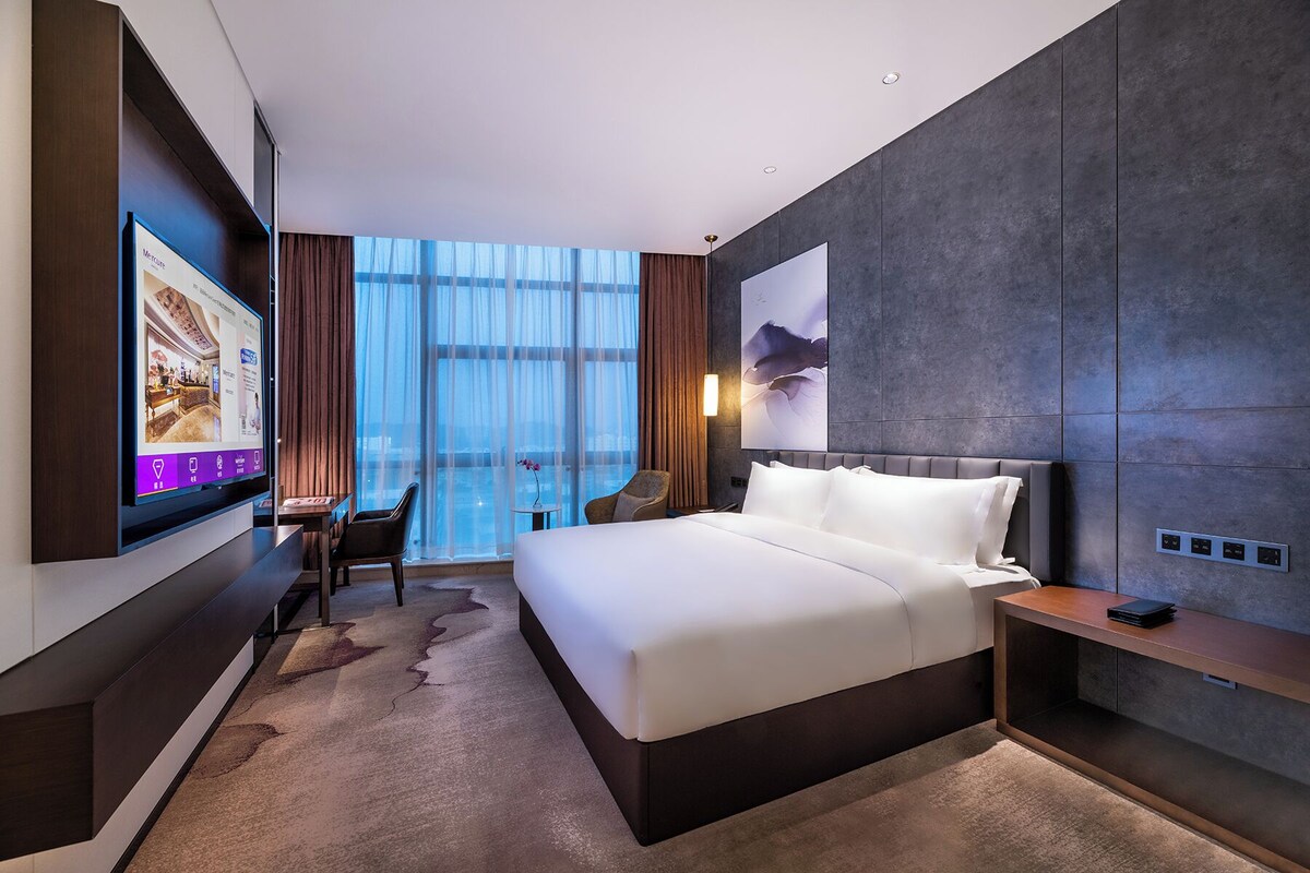 A modern hotel room with a large bed, a desk, a chair, a TV, and a large window with curtains.