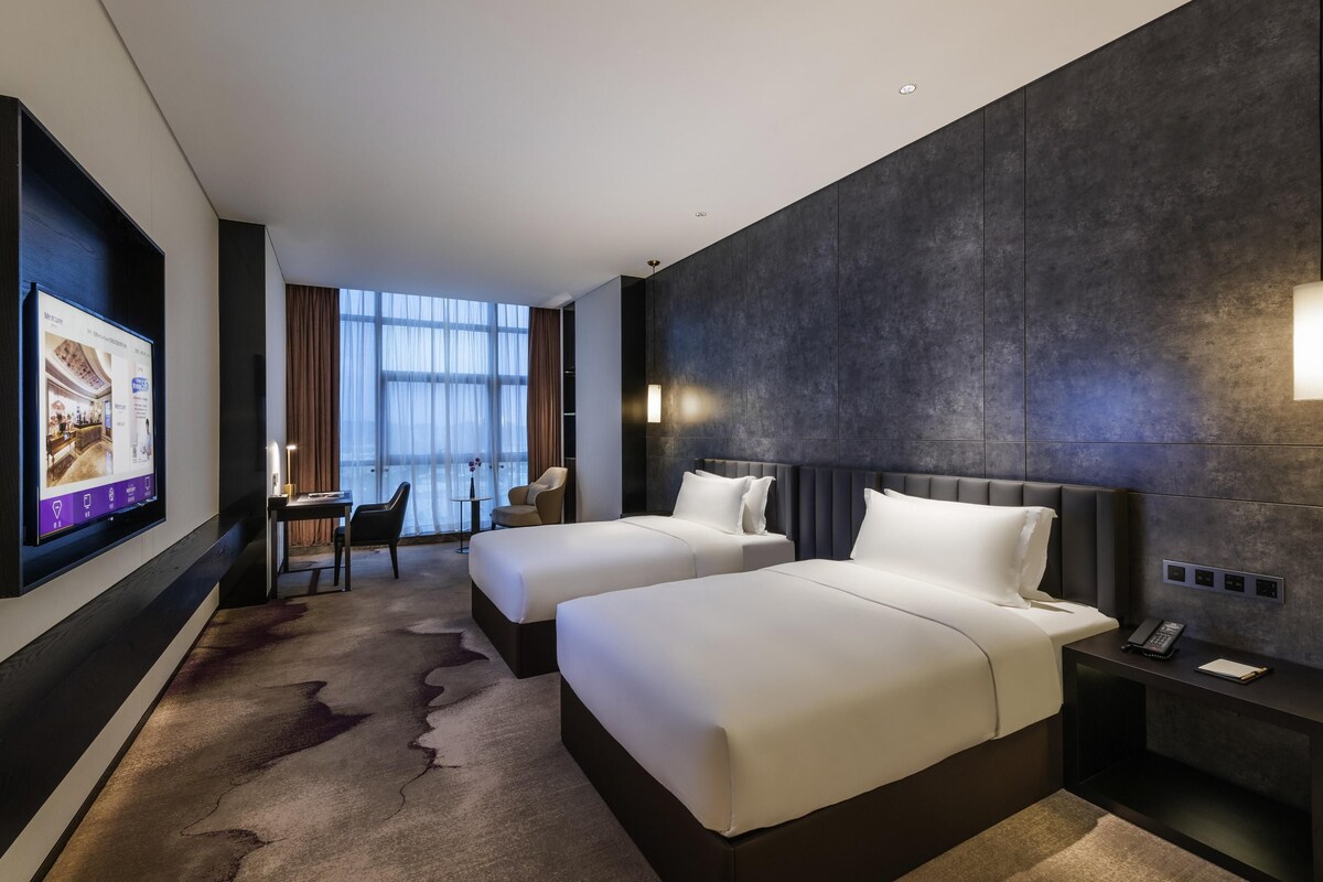 A hotel room with two beds, a desk, a chair, a TV, and a view of the city.