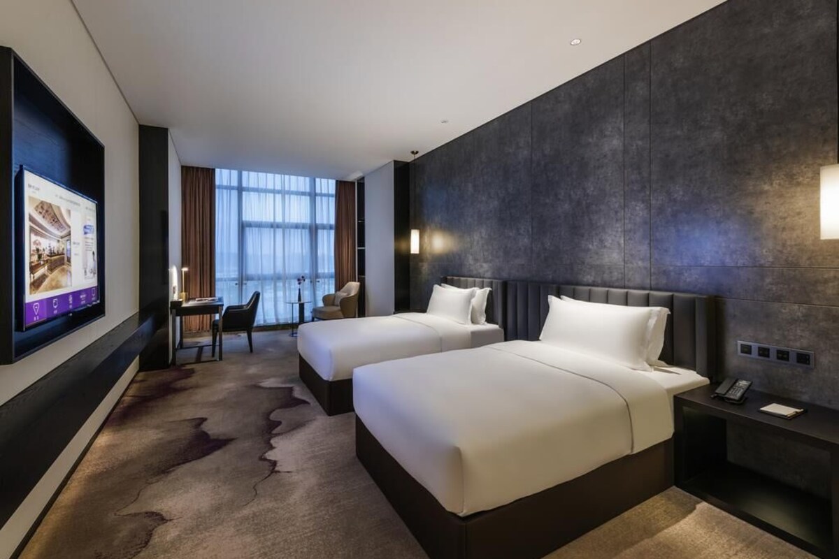 A hotel room with two beds, a TV, a desk, and a view of the city.
