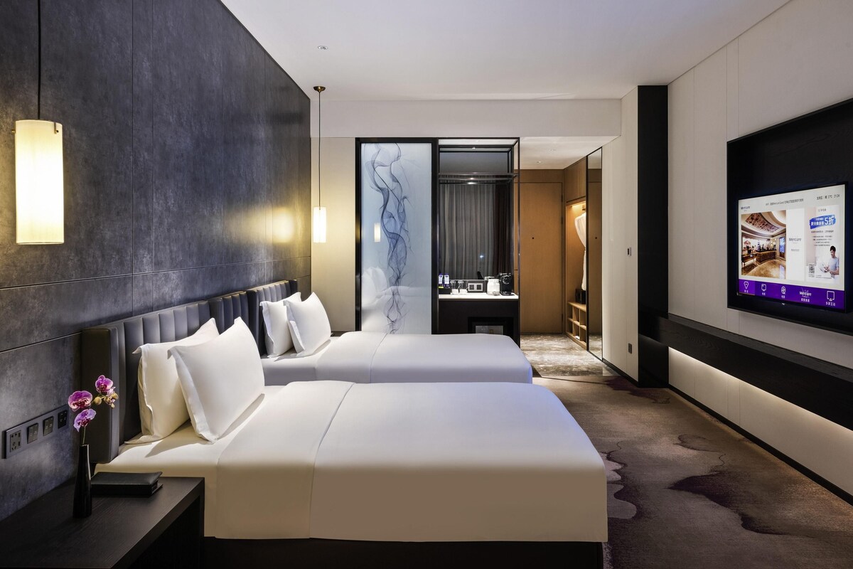 A modern hotel room with two beds, a large mirror, a TV, and a small desk with a vase of flowers.