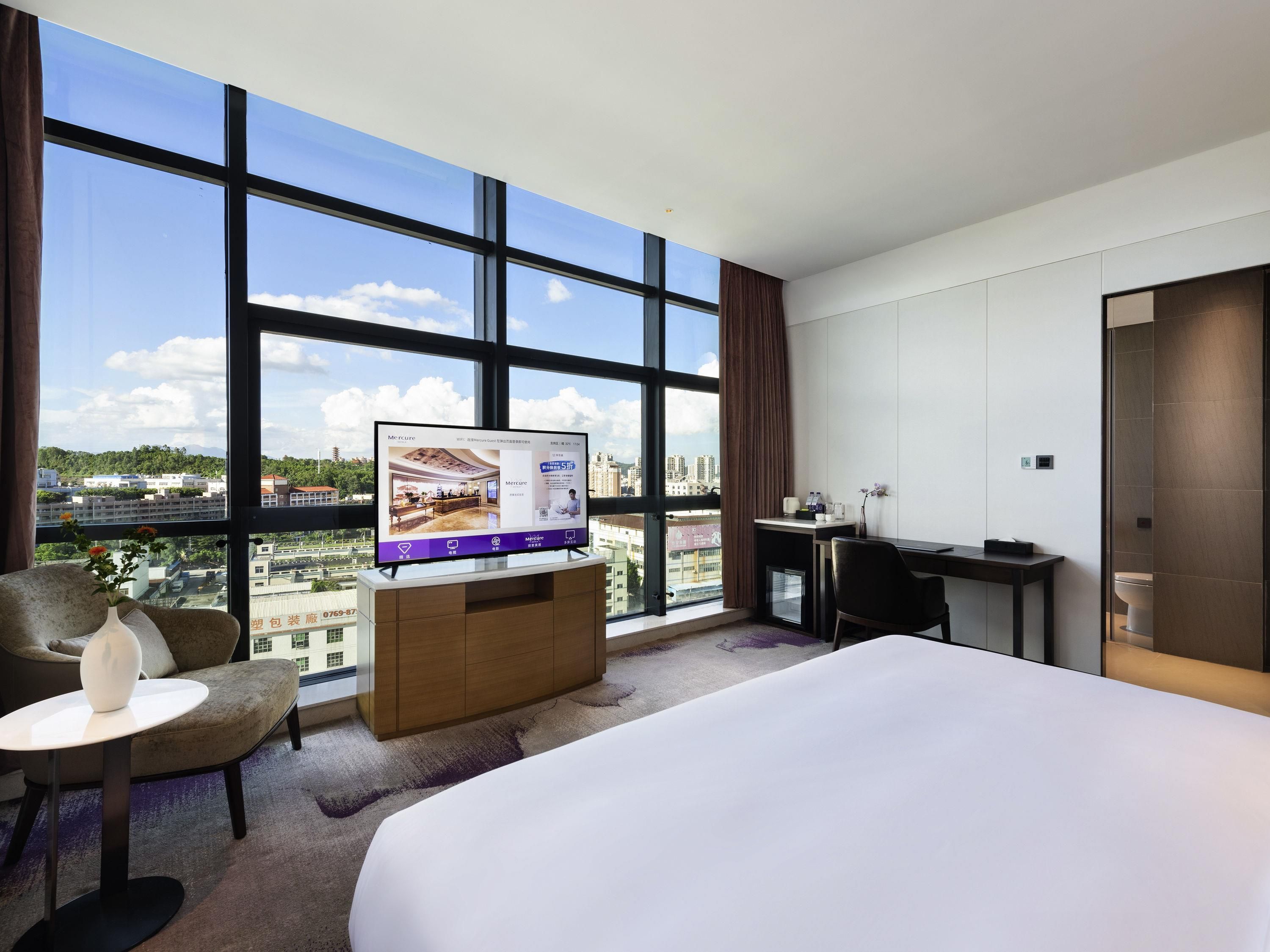 Habitación de hotel con un ventanal grande, una cama, un escritorio, una silla y un televisor que muestra un paisaje urbano.