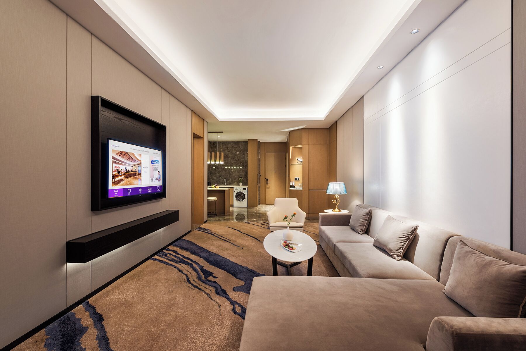 Una habitación de hotel moderna con un televisor de pantalla plana montado en la pared, un sofá, una pequeña mesa redonda y una lámpara.