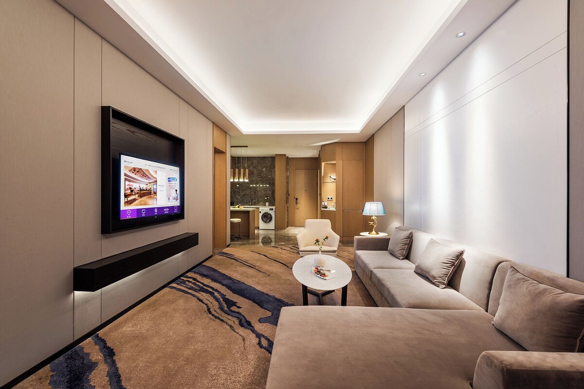 A modern hotel room with a flat-screen TV mounted on the wall, a sofa, a small round table, and a lamp.