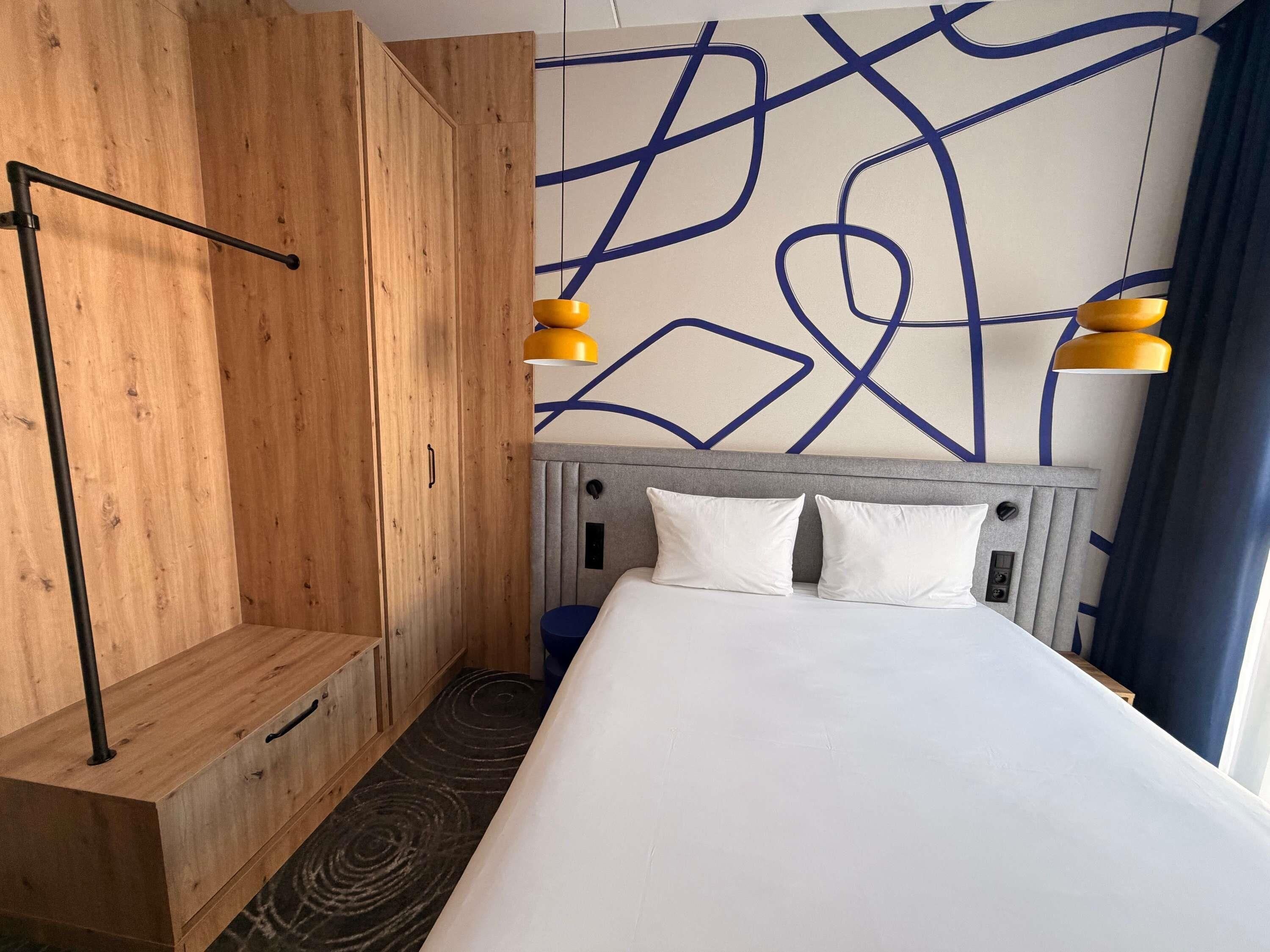 A hotel room with a wooden wardrobe, a bed with white bedding, a headboard with a geometric pattern, and two yellow pendant lights.