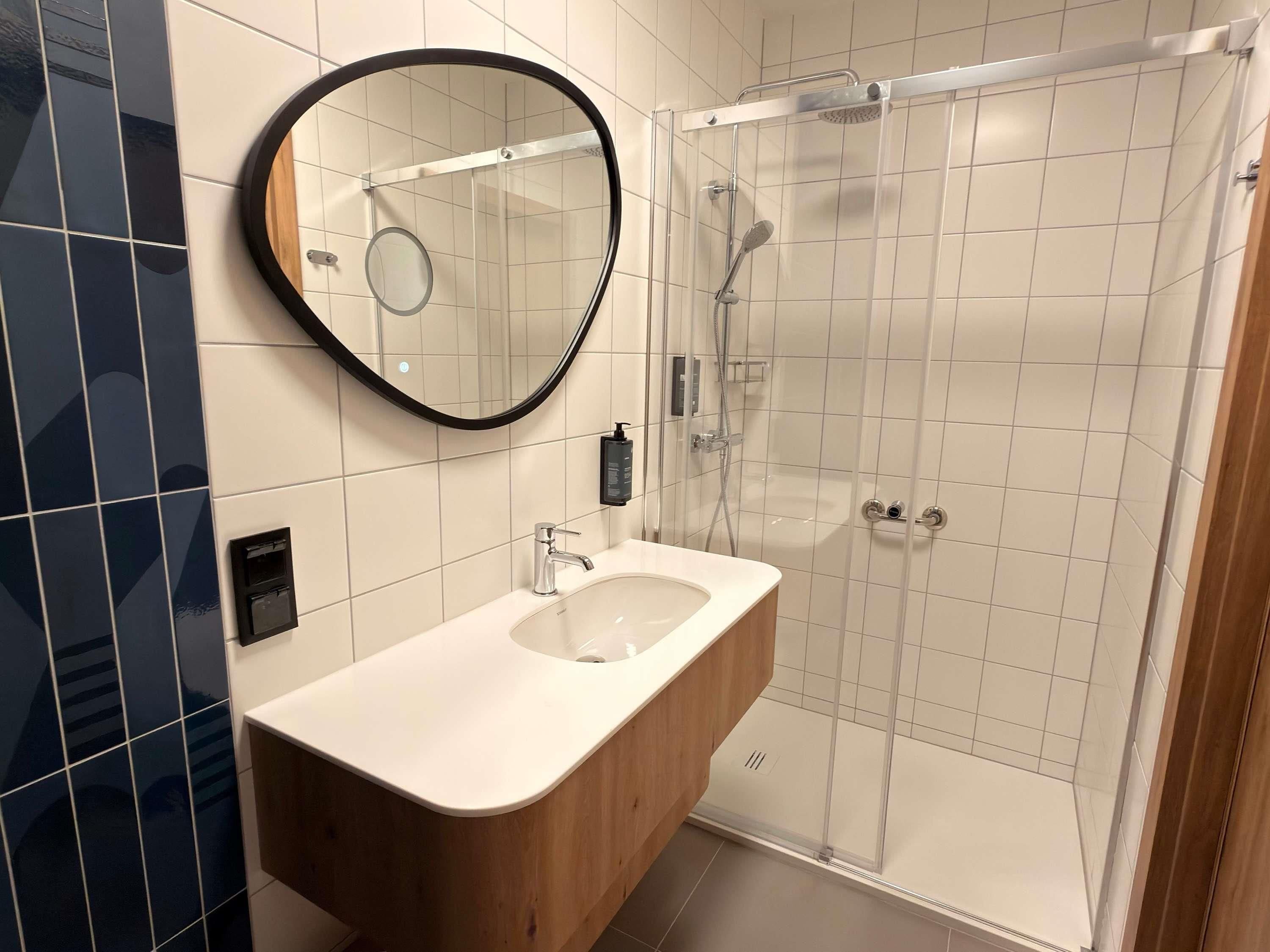 A modern bathroom with a glass-enclosed shower, a wooden vanity, and a round mirror.