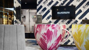 Un vestíbulo moderno con una gran escultura abstracta, un sillón tapizado gris y un televisor que muestra la página web ALLACCOR.COM.