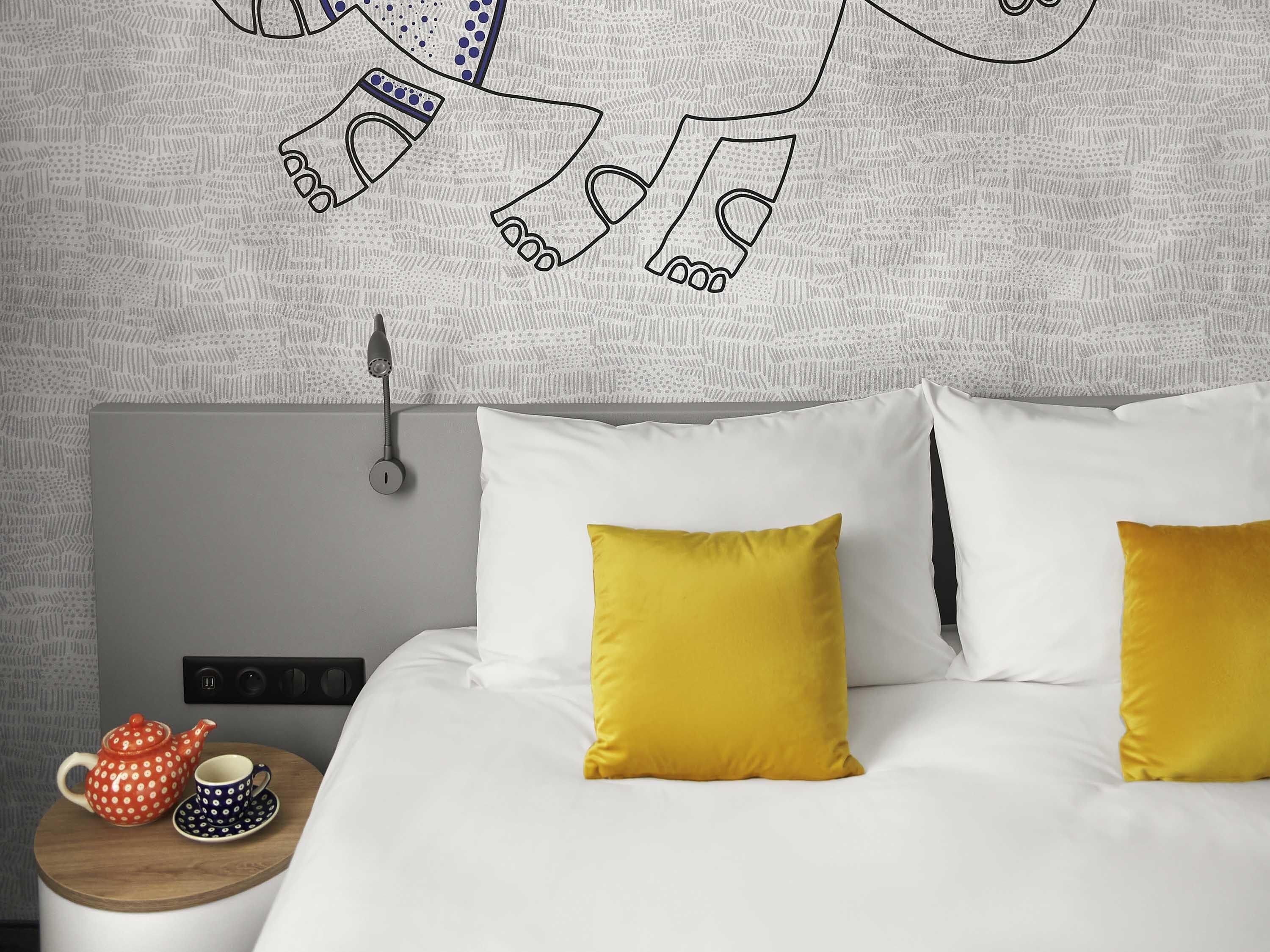 A neatly made bed with two yellow pillows, a bedside table with a teapot and cups, and a wall with a geometric pattern.