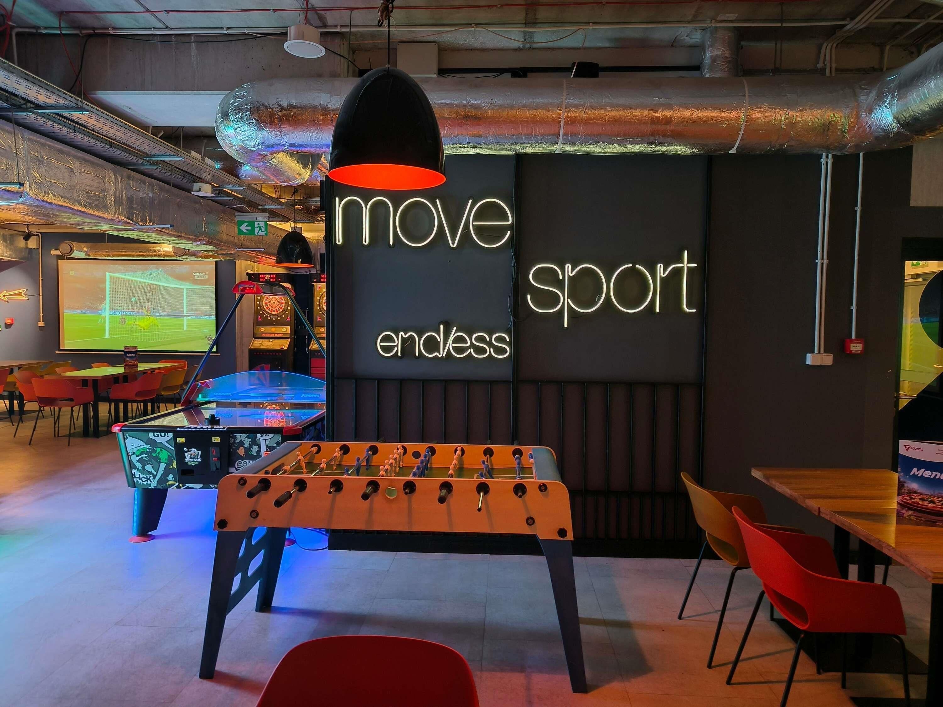 Um espaço de bar com temática esportiva, com mesa de pebolim, letreiros de neon e uma tela exibindo uma partida de futebol.