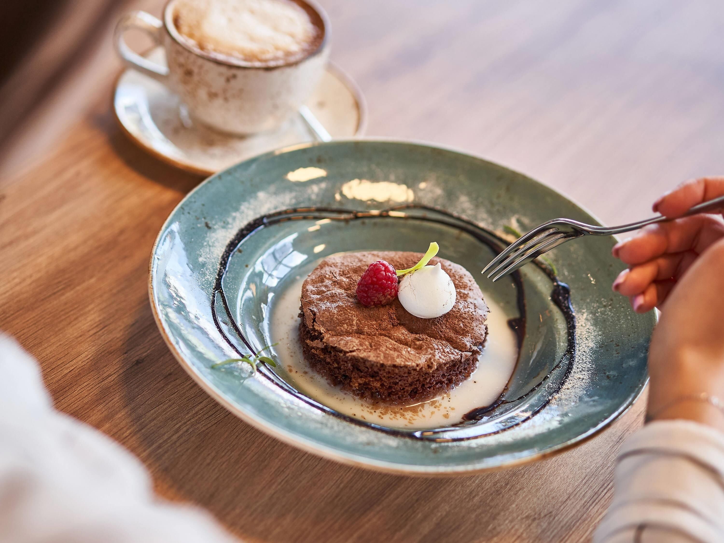 En desserttallrik med en chokladkaka, en hallon och en klick grädde, serverad tillsammans med en kopp kaffe på ett litet fat.