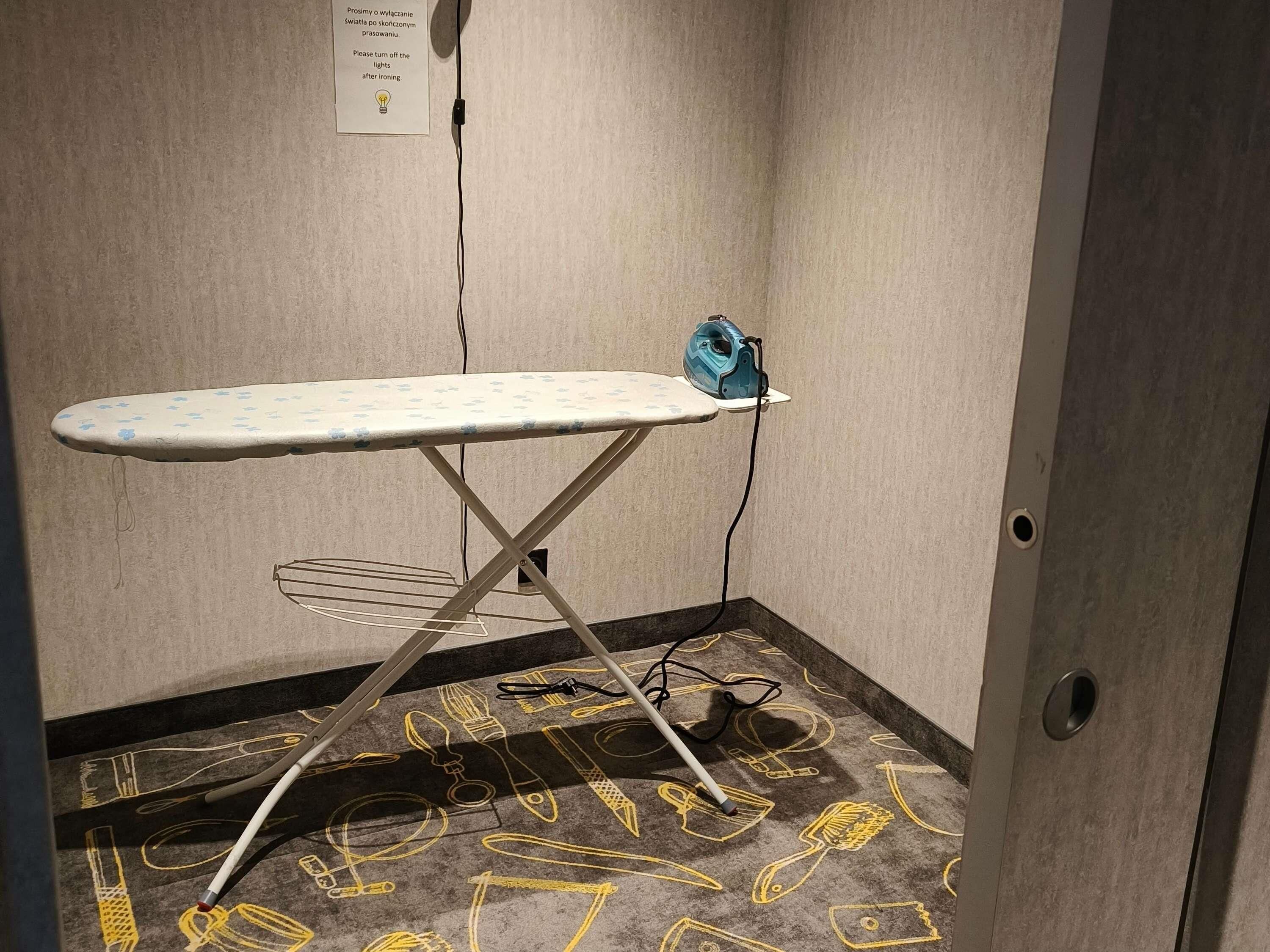 A corner room with a wall-mounted ironing board, a power outlet, and a carpet with yellow patterns.