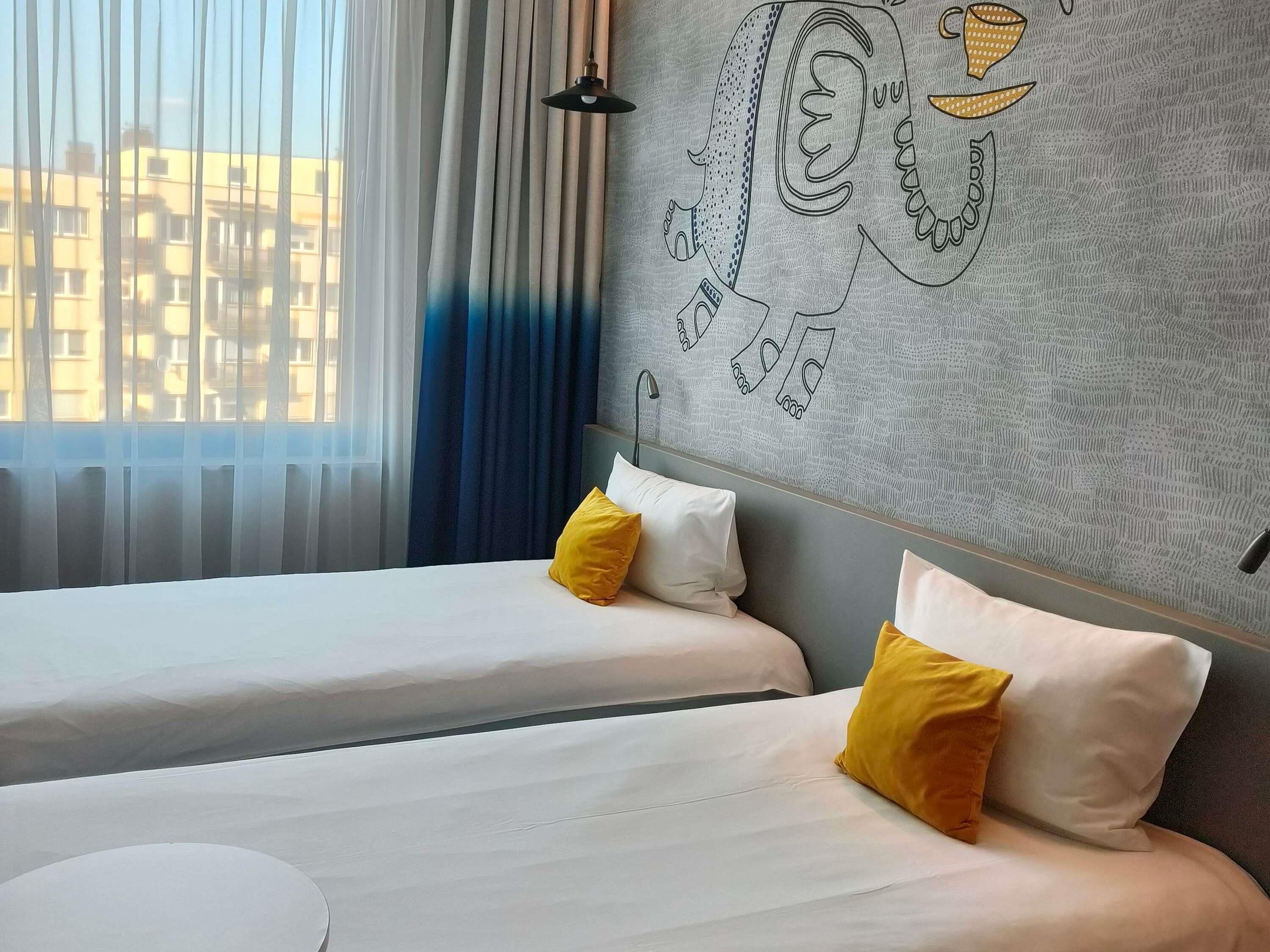 Une chambre d’hôtel avec deux lits, une fenêtre avec des rideaux légers et un mur orné d’un motif géométrique.