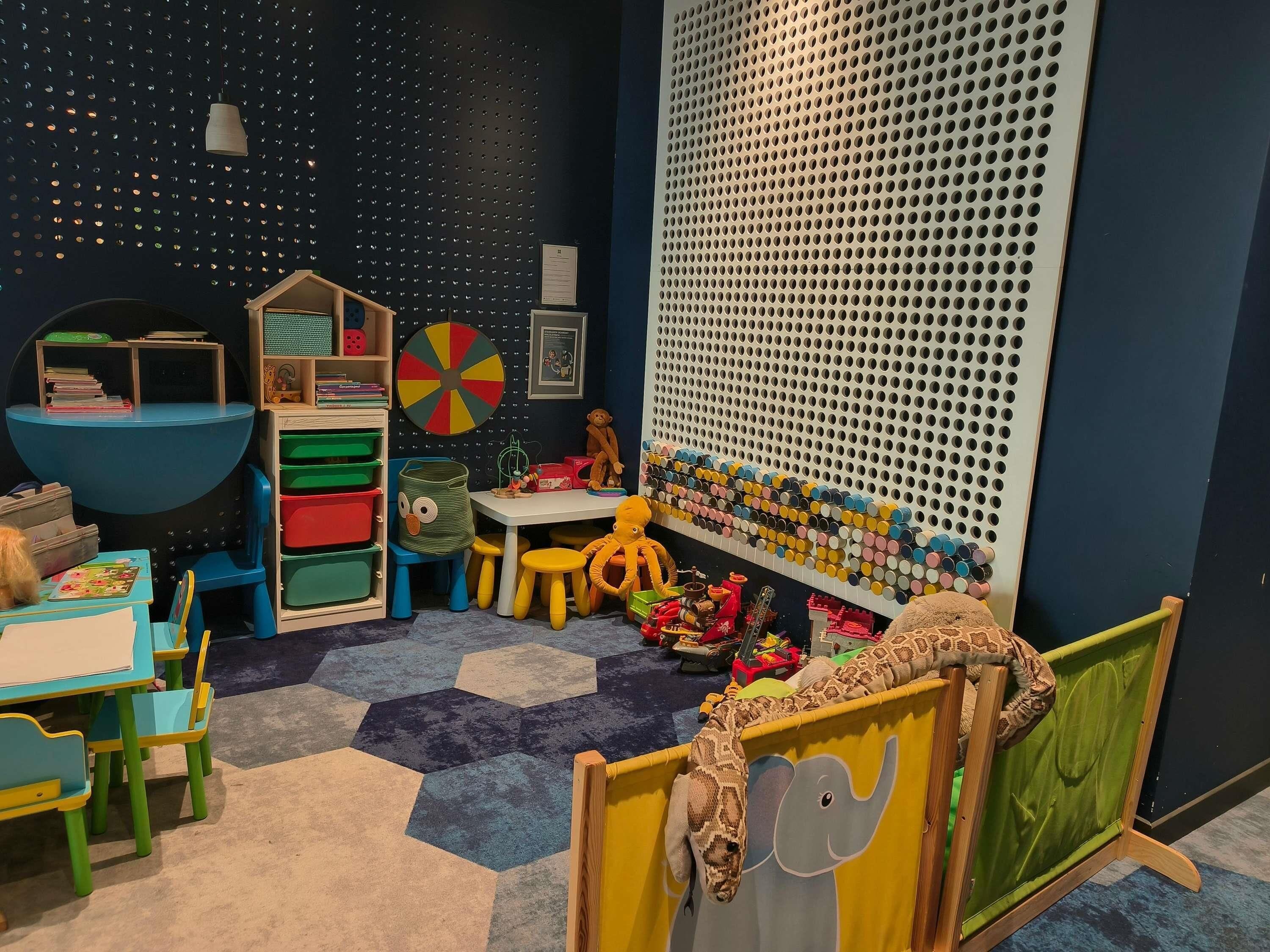 Ein Kinderspielzimmer mit einem sechseckigen Teppich, einem Spieltisch und einer großen Wand mit einem gelochten Design.