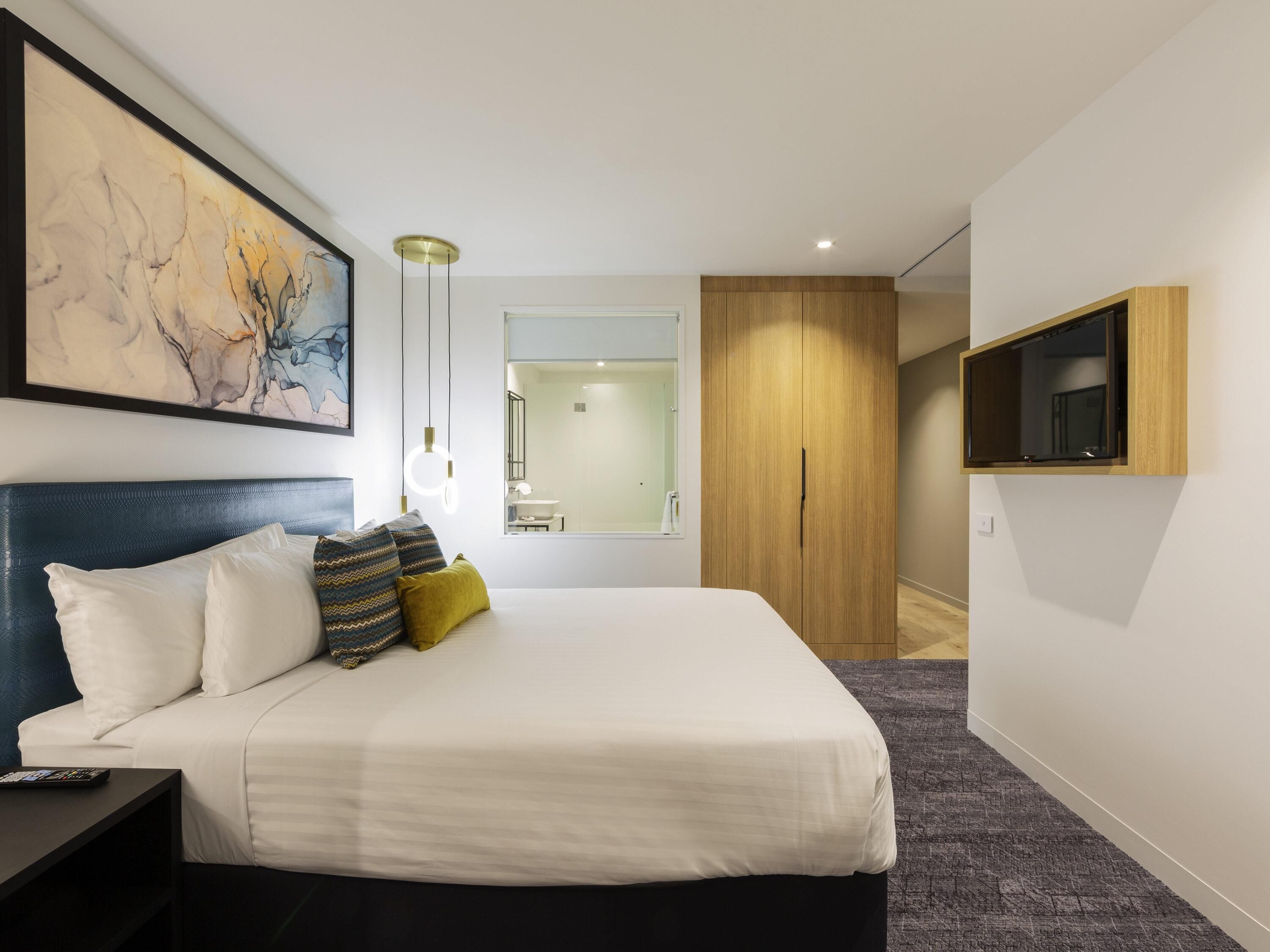 A modern hotel room with a large bed, a TV mounted on the wall, and a painting above the bed.