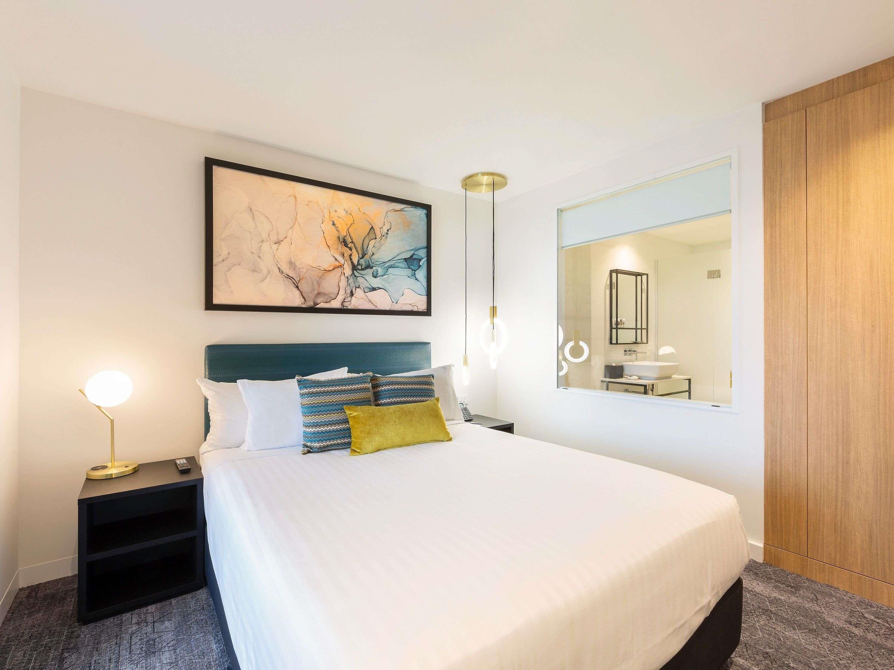 Een hotelkamer met een bed, nachtkastje, lamp, schilderij, spiegel en een kledingkast.