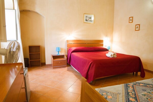 A bedroom with a wooden bed, a nightstand with a lamp, a fan, and a small shelf. - Agriturismo Eloro Pizzuta (Noto)