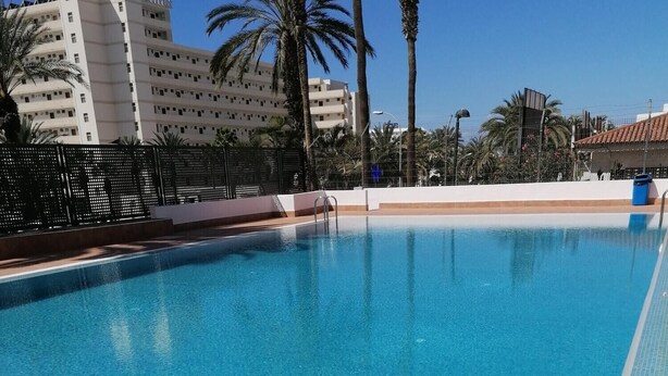 A clear blue swimming pool with a metal handrail, surrounded by palm trees and buildings in the background. - KC home abroad (Playa de las Americas)
