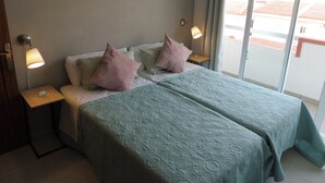 A hotel room with a bed, two bedside lamps, a nightstand, and a view of buildings through a window. - KC home abroad (Playa de las Americas)