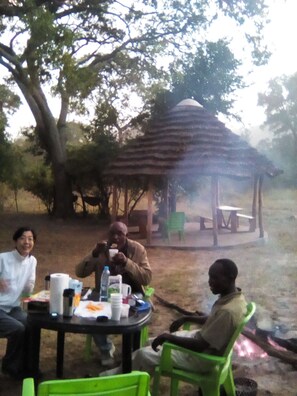 A group of people sitting at an outdoor table with a thatched-roof structure in the background. - Naumba Camp Site (Ngoma)