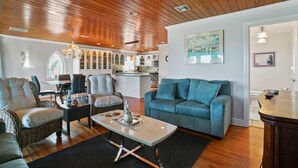 En rummelig stue med træloft, blå sofa, spiseplads med fletstole og et køkken i baggrunden.