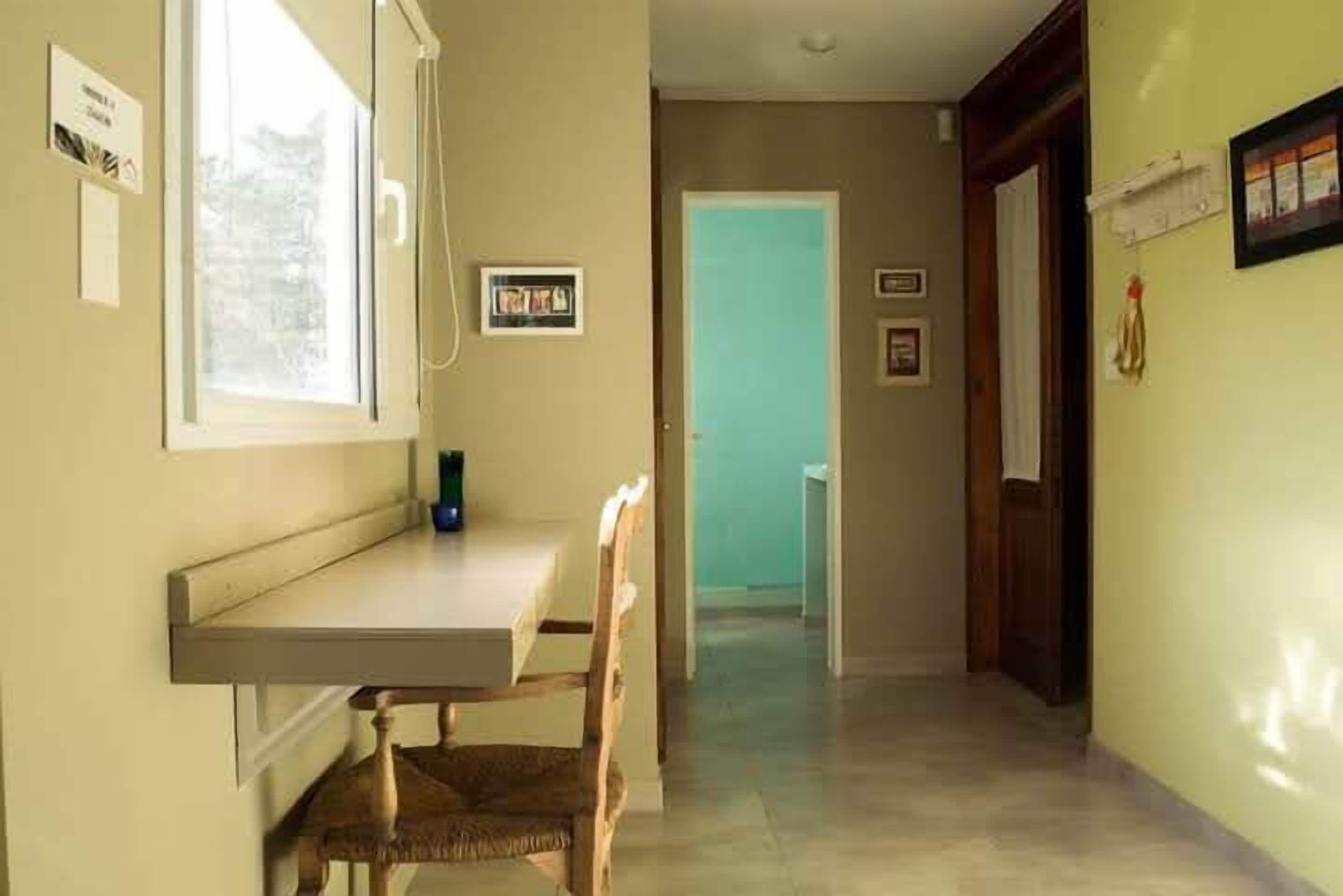 Un couloir étroit, avec une fenêtre sur le côté gauche, une porte au fond et une petite table avec une chaise au centre.