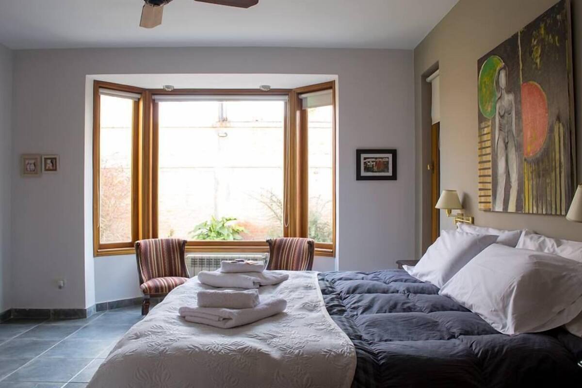 A hotel room with a large bed, a window with a view, a chair, and a painting on the wall.