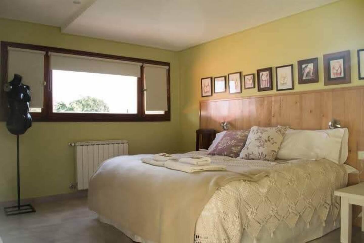 A bedroom with a large bed, a bedside table, a nightstand, a window with blinds, and framed pictures on the wall.