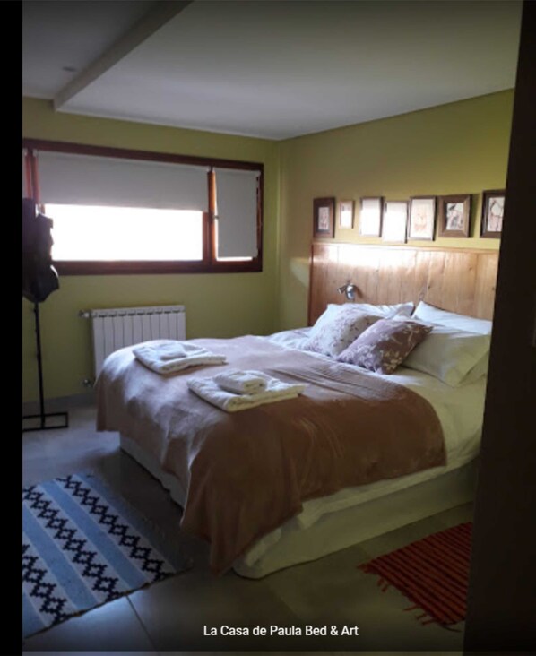 Un dormitorio con cama, ventana, radiador y cuadros en la pared.