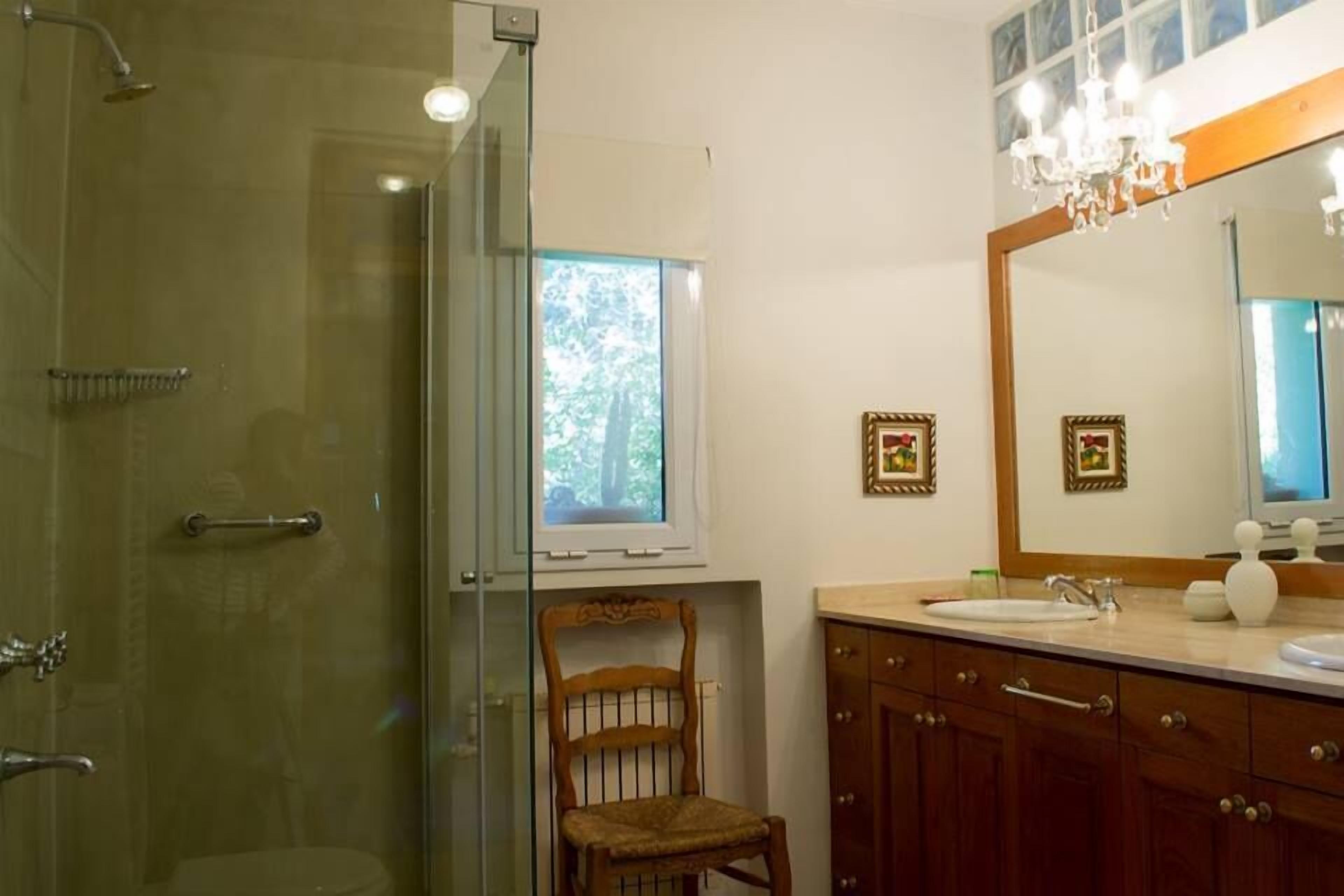 A bathroom with a glass-enclosed shower, a wooden chair, a vanity with a mirror, and two framed pictures on the wall.