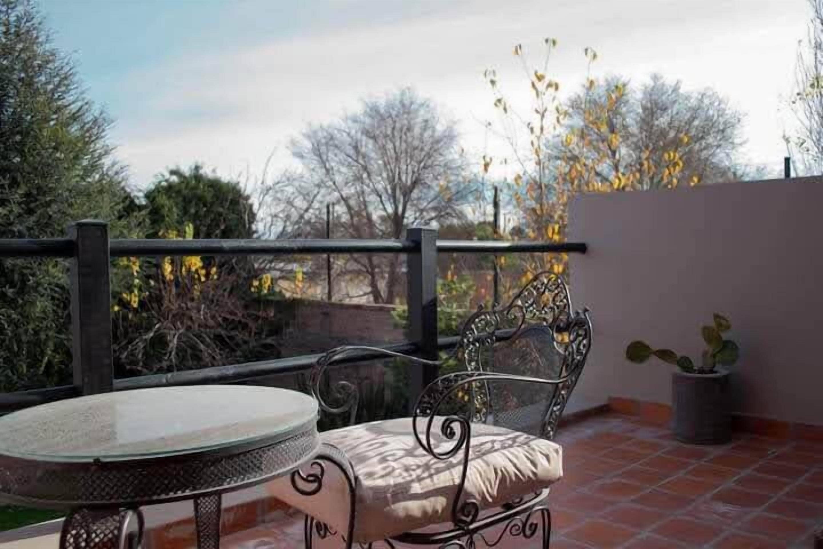 Un balcon avec une table ronde et une chaise en métal, un pot de fleurs et une vue sur des arbres et un bâtiment au loin.