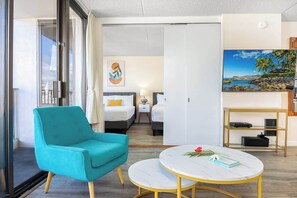 Una habitación de hotel moderna con un sillón azul aguamarina, una mesa redonda de mármol, una cama con ropa de cama blanca, un cuadro de una playa y un estante con dispositivos electrónicos.