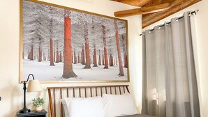 A bedroom with a bed, a nightstand, a lamp, a painting of a snowy forest, and a window with curtains. - 2 Bed Exceptional Fireplace Condo In the Heart of Town On Shuttle Route (Ludlow)